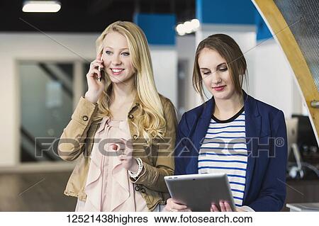 Stock Photograph - Two beautiful young millennial business women using technology in the workplace; Sherwood Park, Alberta, Canada. Fotosearch