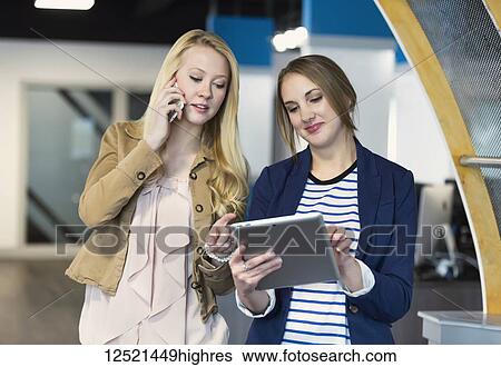 Stock Photograph - Two beautiful young millennial business women using technology in the workplace; Sherwood Park, Alberta, Canada. Fotosearch