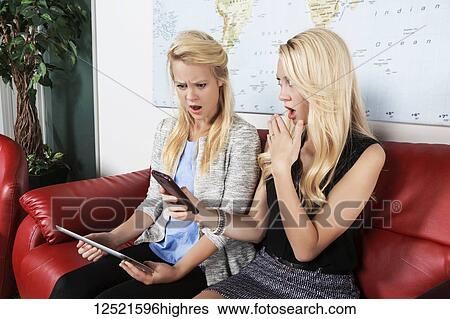 Two beautiful young millennial business women sitting in a workplace using their technology with shocked looks on their faces as they read something on social media; Sherwood Park, Alberta, Canada View Large Photo Image Stock Photograph - Two beautiful young millennial business women sitting in a workplace using their technology with shocked looks on their faces as they read something on social media; Sherwood Park, Alberta, Canada. Fotosearch