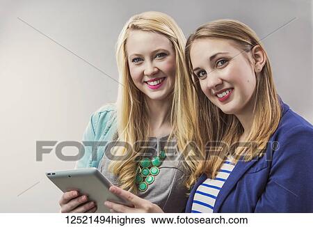 Stock Photograph - Two beautiful young women who are millennial business professionals working together using a tablet and posing for the camera at their workplace; Sherwood Park, Alberta, Canada. Fotosearch