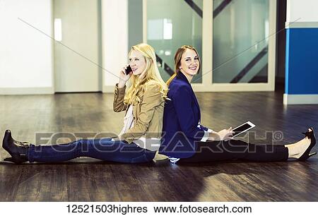 Stock Photograph - Two beautiful young women who are millennial business professionals sitting back to back on the floor of their workplace using technology; Sherwood Park, Alberta, Canada. Fotosearch