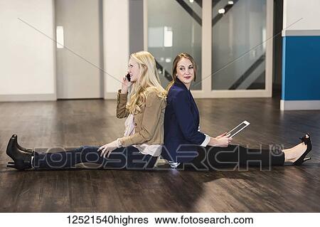 Stock Photograph - Two beautiful young women who are millennial business professionals sitting back to back on the floor of their workplace using technology; Sherwood Park, Alberta, Canada. Fotosearch
