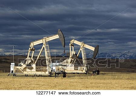 Two dramatically sun lit pumpjacks in a field with dark clouds in the sky and shadowed mountains in the background; Alberta, Canada View Large Photo Image Stock Photo - Two dramatically sun lit pumpjacks in a field with dark clouds in the sky and shadowed mountains in the background; Alberta, Canada. Fotosearch