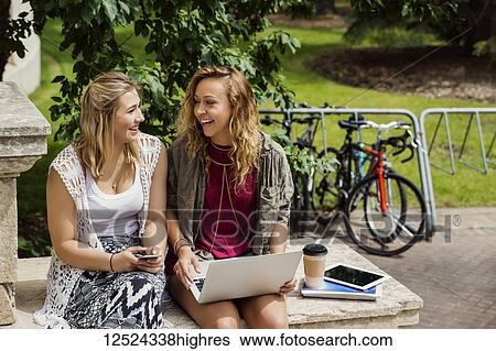 Stock Photograph - Two female university students laughing and talking together while using their technology outside on the campus; Edmonton, Alberta, Canada. Fotosearch