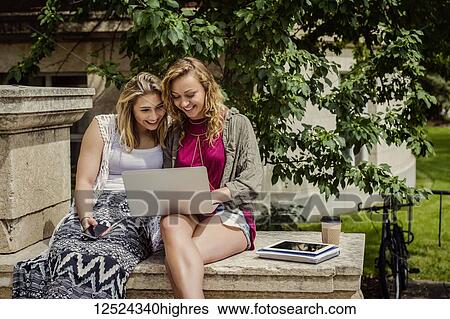Stock Photograph - Two female university students sitting together and looking at a laptop computer outside on the steps of a campus building; Edmonton, Alberta, Canada. Fotosearch