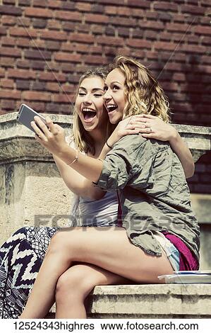 Stock Photograph - Two female university students sitting together on the campus, being silly and taking a self-portrait with their smart phone; Edmonton, Alberta, Canada. Fotosearch
