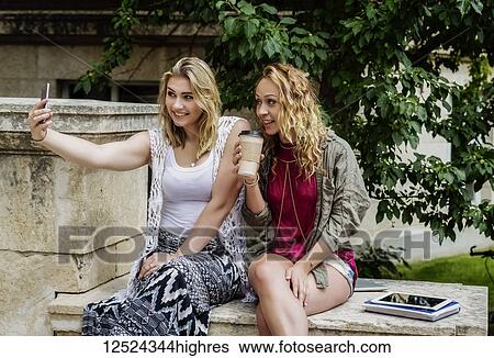 Stock Photograph - Two female university students sitting together on the campus taking a self-portrait with their smart phone; Edmonton, Alberta, Canada. Fotosearch