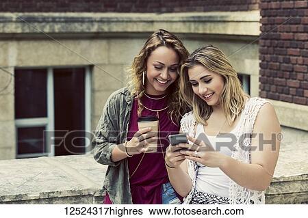 Stock Photograph - Two female university students standing together on the campus looking at a smart phone; Edmonton, Alberta, Canada. Fotosearch