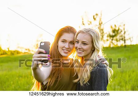 Stock Photograph - Two friends standing in a park taking a self-portrait with a smart phone at dusk; Edmonton, Alberta, Canada. Fotosearch
