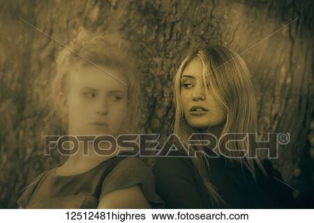 Stock Photograph - Two teenage girls with blond hair standing back to back beside a tree trunk looking serious, as if in conflict; Connecticut, United States of America. Fotosearch