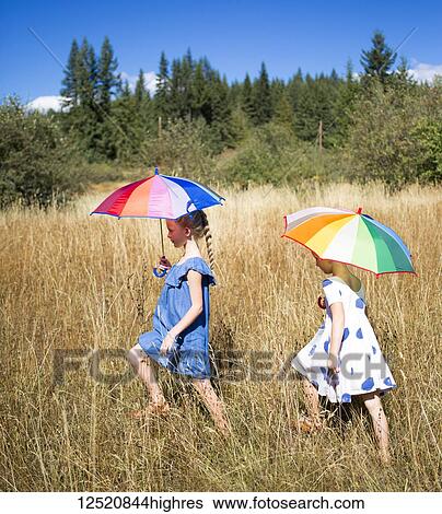 Two young girls wearing sundresses walk through a field with tall grass holding umbrellas; Salmon Arm, British Columbia, Canada View Large Photo Image Stock Photograph - Two young girls wearing sundresses walk through a field with tall grass holding umbrellas; Salmon Arm, British Columbia, Canada. Fotosearch