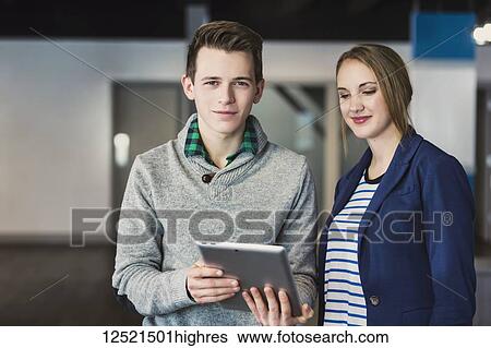 Stock Photograph - Two young millennial business professionals using technology in the workplace and one looking at the camera; Sherwood Park, Alberta, Canada. Fotosearch
