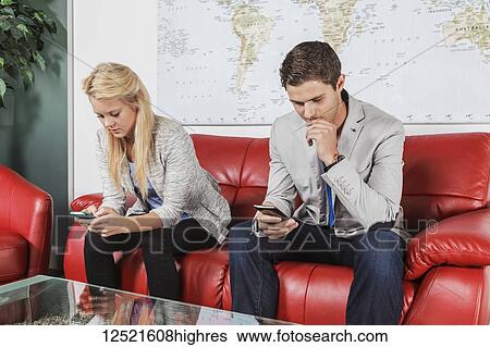 Stock Photograph - Two young millennial business professionals in the lobby of their workplace using their smart phones while taking a break; Sherwood Park, Alberta, Canada. Fotosearch