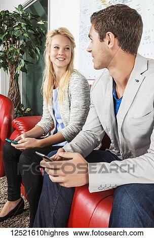 Stock Photograph - Two young millennial business professionals in the lobby of their workplace using their smart phones while taking a break; Sherwood Park, Alberta, Canada. Fotosearch