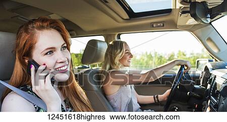 Stock Photograph - Two young women driving in a car on a road trip while one talks on a smart phone; Edmonton, Alberta, Canada. Fotosearch