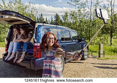 Two young women on a road trip sit in the back of a vehicle talking and laughing together while a friend takes a selfie with a selfie stick; Edmonton, Alberta, Canada View Large Photo Image Stock Photograph - Two young women on a road trip sit in the back of a vehicle talking and laughing together while a friend takes a selfie with a selfie stick; Edmonton, Alberta, Canada. Fotosearch