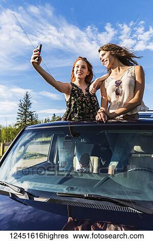 Stock Photograph - Two young women on a road trip stand up in the sunroof of a vehicle taking a self-portrait with a smart phone; Edmonton, Alberta, Canada. Fotosearch