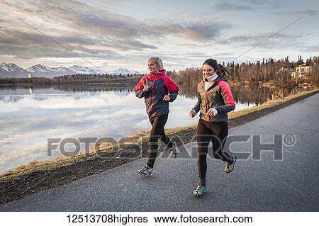 Stock Photograph - Two young women running on a trail at the water's edge with mountains in the distance; Anchorage, Alaska, United States of America. Fotosearch