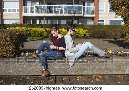 Stock Photograph - Two young women sit back to back using their smart phones; New Westminster, British Columbia, Canada. Fotosearch