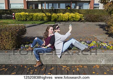 Stock Photograph - Two young women sit back to back using their smart phones and being silly; New Westminster, British Columbia, Canada. Fotosearch