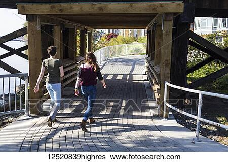 Stock Photograph - Two young women walk together down a path under a bridge in a park; New Westminster, British Columbia, Canada. Fotosearch