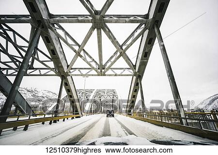 Stock Photograph - Vehicles traveling on a snow covered road over a bridge with a view from the front of a vehicle; Nelson, British Columbia, Canada. Fotosearch