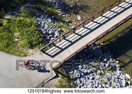 Stock Photograph - View from directly above a cyclist in a red shirt going down a path and onto a bridge over a creek; Calgary, Alberta, Canada. Fotosearch