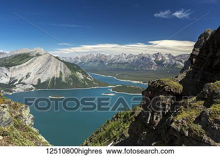 Stock Photograph - View from the top of mountain ridge looking down on colourful alpine lake and mountain range in the distance with blue sky and clouds; Kananaskis Country, Alberta, Canada. Fotosearch