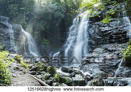 Stock Photograph - Waterfall over rugged rock cliffs and beams of sunlight streaming through the mist; North Carolina, United States of America. Fotosearch