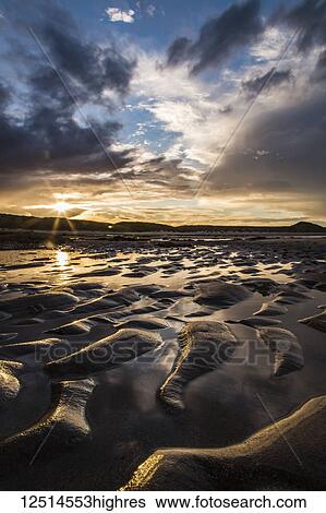 Stock Photograph - Wet ripples in the sand on a beach at sunset with a sunburst over silhouetted hills in the distance; Embleton, Northumberland, England. Fotosearch