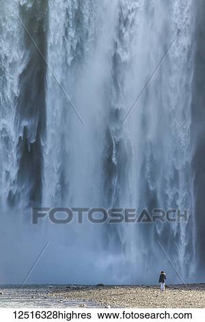 Woman standing at base of Skogafoss waterfall and looking at the powerful flowing water; Iceland View Large Photo Image Stock Photograph - Woman standing at base of Skogafoss waterfall and looking at the powerful flowing water; Iceland. Fotosearch