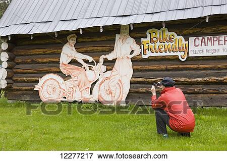 Woman tourist photographs a log cabin and motorcycle sign at the Tetsa River Lodge, British Columbia, Canada, Summer View Large Photo Image Stock Photo - Woman tourist photographs a log cabin and motorcycle sign at the Tetsa River Lodge, British Columbia, Canada, Summer. Fotosearch