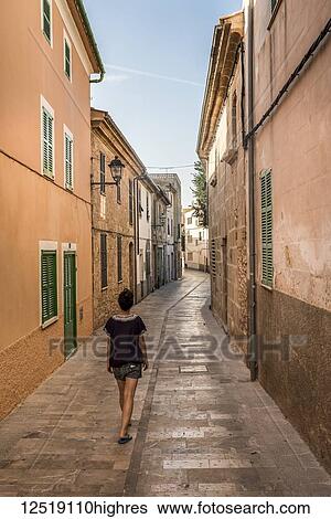 Stock Photograph - Woman walking down a narrow street; Alcudia, Mallorca, Balearic Islands, Spain. Fotosearch