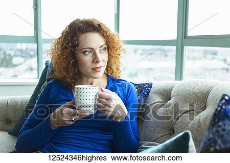 Stock Photograph - Woman with red, curly hair sitting with a mug by a window; Surrey, British Columbia, Canada. Fotosearch