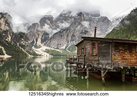 Stock Photograph - Wooden lake boat house with misty clouded mountain in the background and lake reflection; Sesto, Bolzano, Italy. Fotosearch