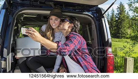 Stock Photograph - Young couple at their vehicle with the back open looking at a laptop computer and taking a self-portrait with a smart phone; Edmonton, Alberta, Canada. Fotosearch