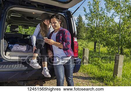 Stock Photograph - Young couple at their vehicle with the back open looking at a cell phone; Edmonton, Alberta, Canada. Fotosearch