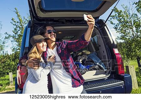 Young couple at their vehicle with the back open taking a self-portrait with their cell phone; Edmonton, Alberta, Canada View Large Photo Image Stock Photograph - Young couple at their vehicle with the back open taking a self-portrait with their cell phone; Edmonton, Alberta, Canada. Fotosearch
