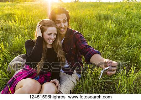 Stock Photograph - Young couple in a park posing for a self-portrait with their cell phone; Edmonton, Alberta, Canada. Fotosearch