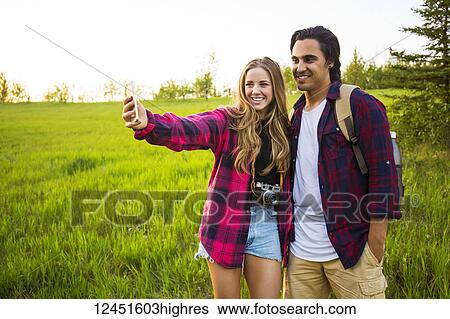Stock Photograph - Young couple in a park posing for a self-portrait with their cell phone; Edmonton, Alberta, Canada. Fotosearch