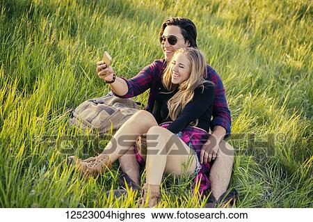 Stock Photograph - Young couple sit on the grass in a park posing for a self-portrait with their cell phone; Edmonton, Alberta, Canada. Fotosearch