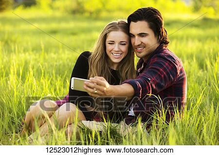 Stock Photograph - Young couple sit on the grass in a park posing for a self-portrait with their cell phone; Edmonton, Alberta, Canada. Fotosearch