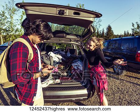 Stock Photograph - Young couple standing at the back of their packed vehicle with back door open and the young man in using his cell phone while the young woman waits impatiently; Edmonton, Alberta, Canada. Fotosearch