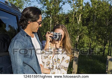 Young couple with camera standing outside their vehicle, the young woman taking a picture in the distance; Edmonton, Alberta, Canada View Large Photo Image Stock Photograph - Young couple with camera standing outside their vehicle, the young woman taking a picture in the distance; Edmonton, Alberta, Canada. Fotosearch