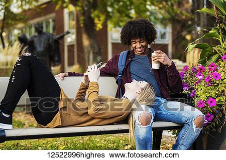 Stock Photograph - Young dating couple who are university students relaxing on a bench on campus and the woman is checking her smart phone while laying her head on her boyfriend's lap; Edmonton, Alberta, Canada. Fotosearch