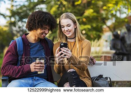 Stock Photograph - Young dating couple who are university students sitting on a bench on campus and looking at social media on a smart phone; Edmonton, Alberta, Canada. Fotosearch