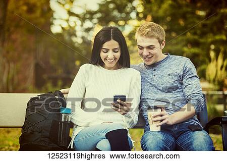 Stock Photograph - Young dating couple who are university students sitting on a bench on campus and looking at social media on a smart phone; Edmonton, Alberta, Canada. Fotosearch
