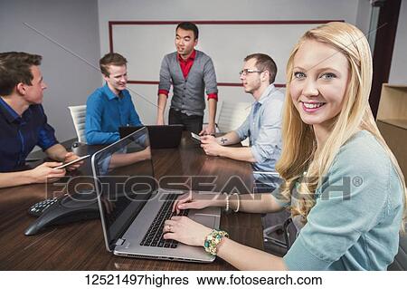 Young millennial business woman posing for the camera while a co-worker is speaking to a group of her piers in a conference room; Sherwood Park, Alberta, Canada View Large Photo Image Stock Photograph - Young millennial business woman posing for the camera while a co-worker is speaking to a group of her piers in a conference room; Sherwood Park, Alberta, Canada. Fotosearch