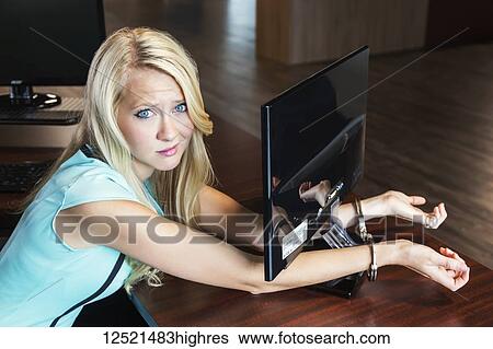Stock Photograph - Young millennial business woman handcuffed to her computer monitor in the workplace; Sherwood Park, Alberta, Canada. Fotosearch