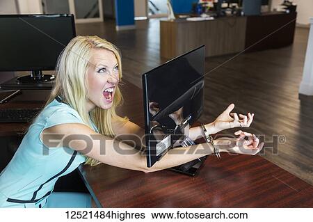 Stock Photograph - Young millennial business woman handcuffed to her computer monitor in the workplace; Sherwood Park, Alberta, Canada. Fotosearch